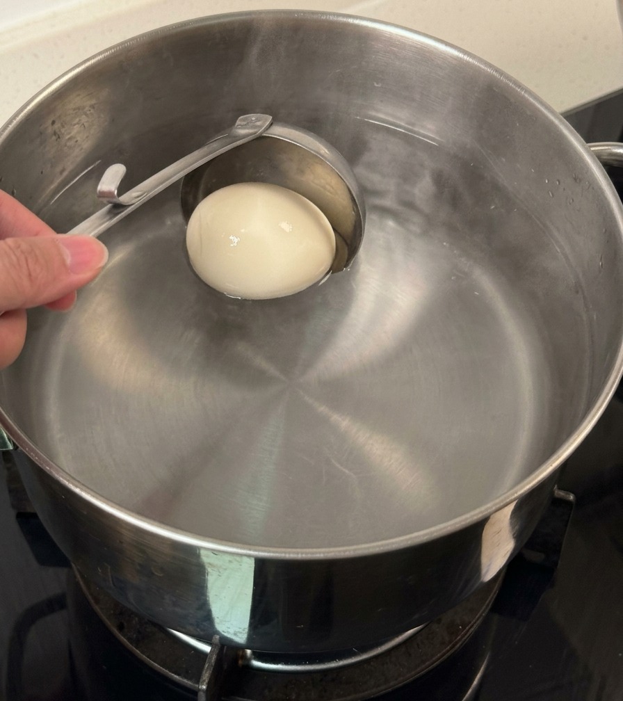 Un mestolo di metallo cala delicatamente un uovo intero con guscio bianco in una pentola dacqua.