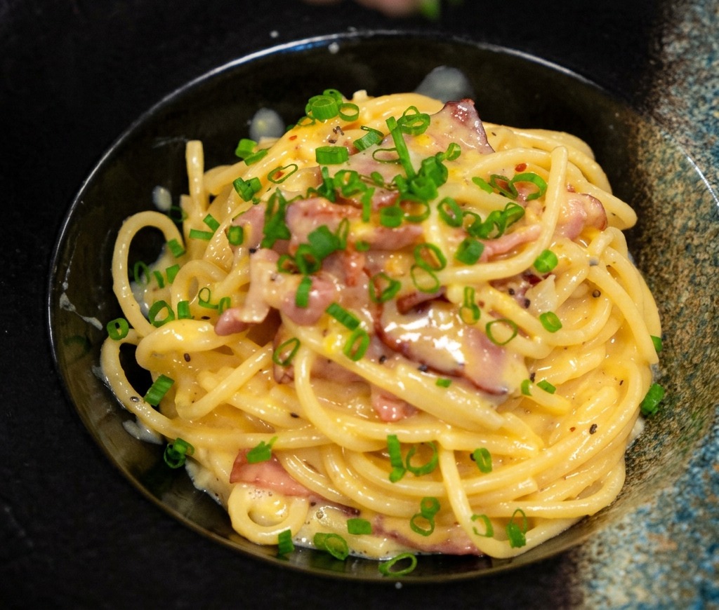 Dita che spolverano cipollotti tritati su un piatto di pasta cremosa alla pancetta.