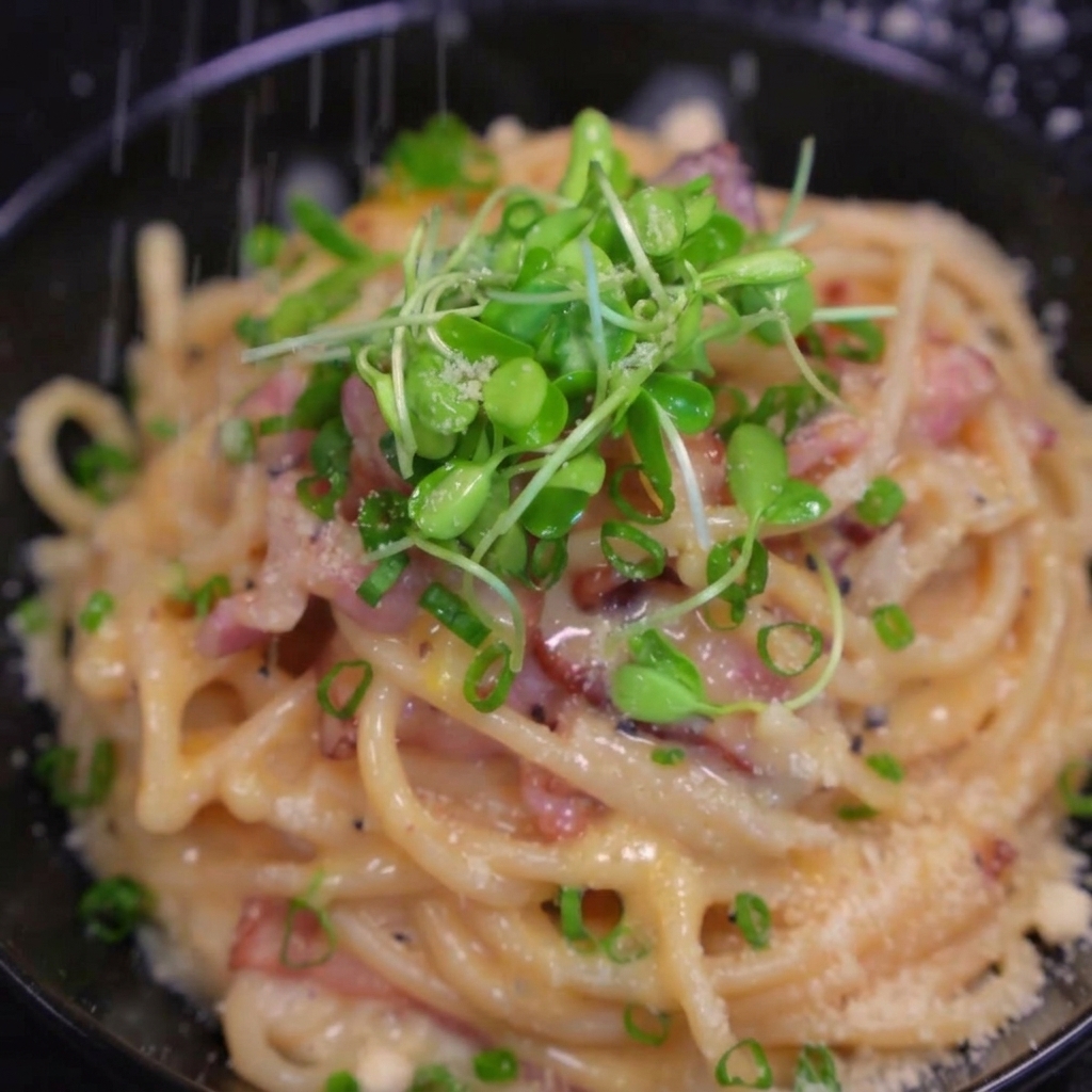 Formaggio fresco grattugiato sopra un piatto di pasta alla pancetta guarnito con cipollotti.