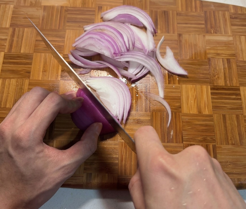 Mani che affettano una cipolla rossa a strisce sottili su un tagliere di bambù con un coltello da chef affilato.