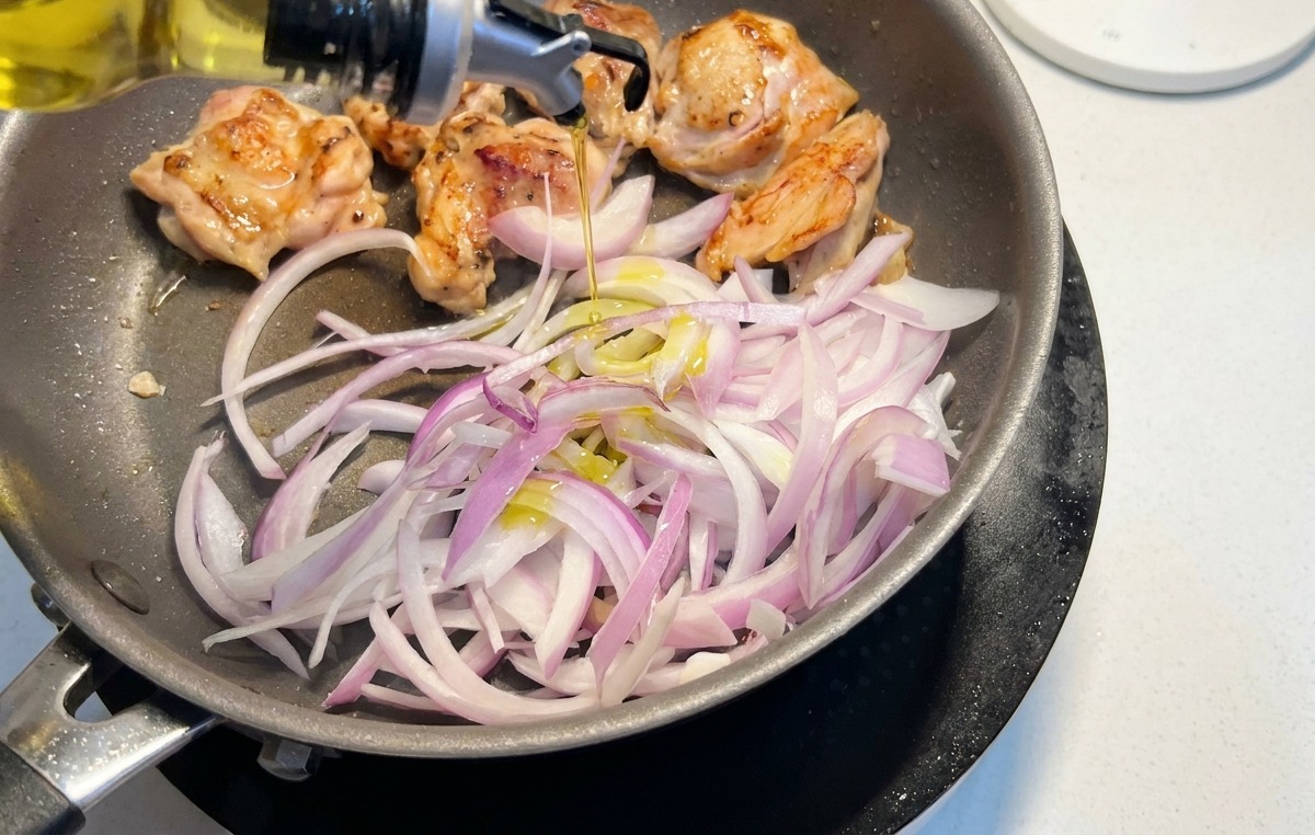 Cipolle rosse affettate aggiunte in padella con le sovracosce di pollo che dorano mentre viene versato un filo d olio.