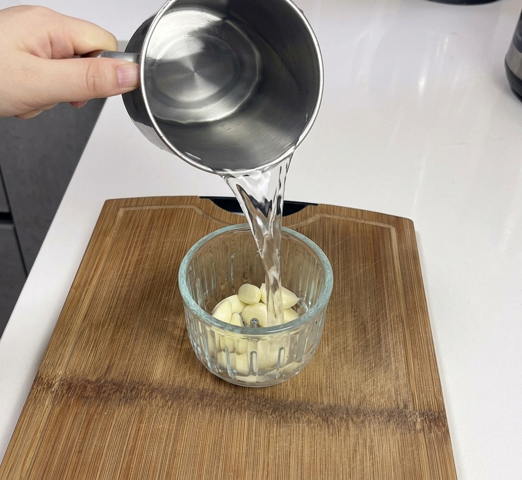 Un boccale di metallo versa acqua in un frullatore di vetro riempito con spicchi daglio fresco.