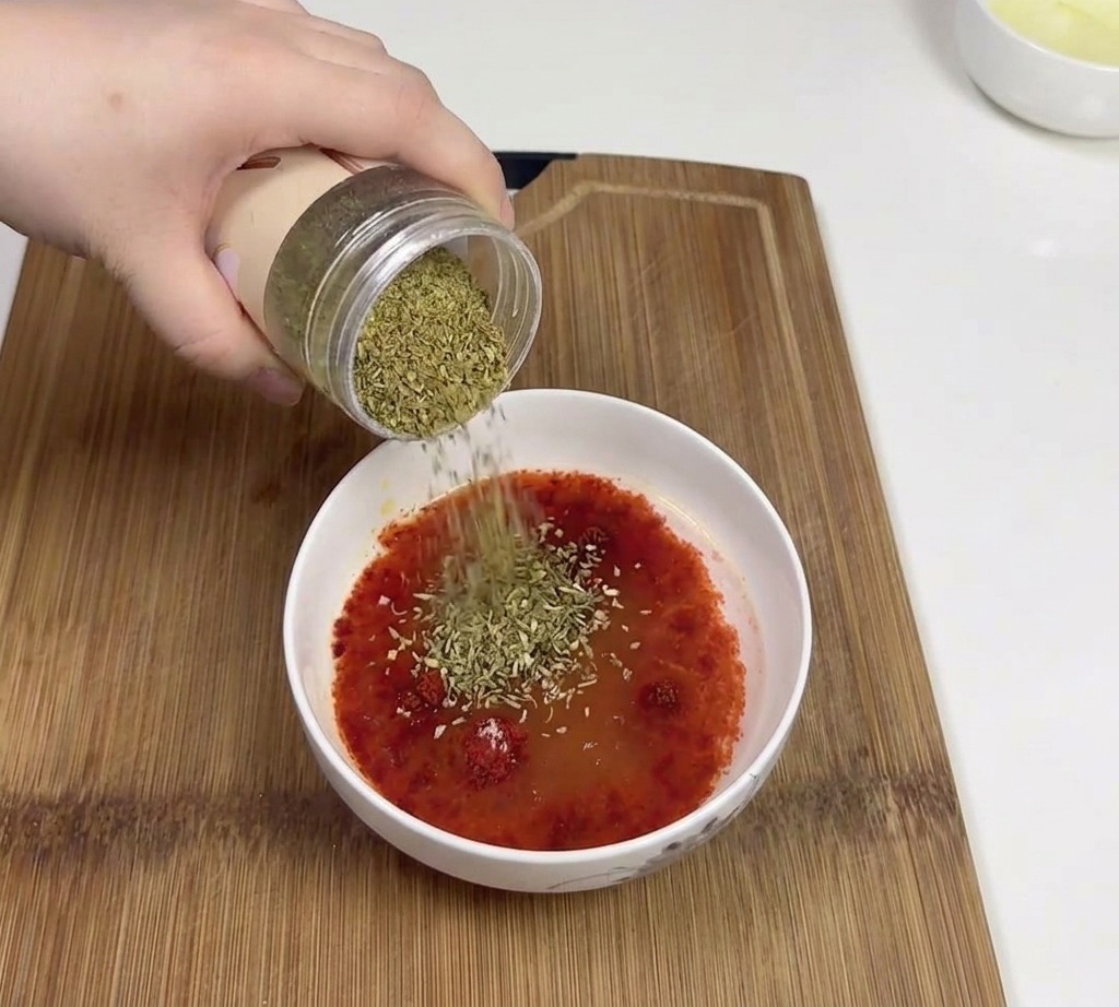 Una mano versa del cumino in polvere da un barattolo in una ciotola contenente una base di salsa al peperoncino rosso.