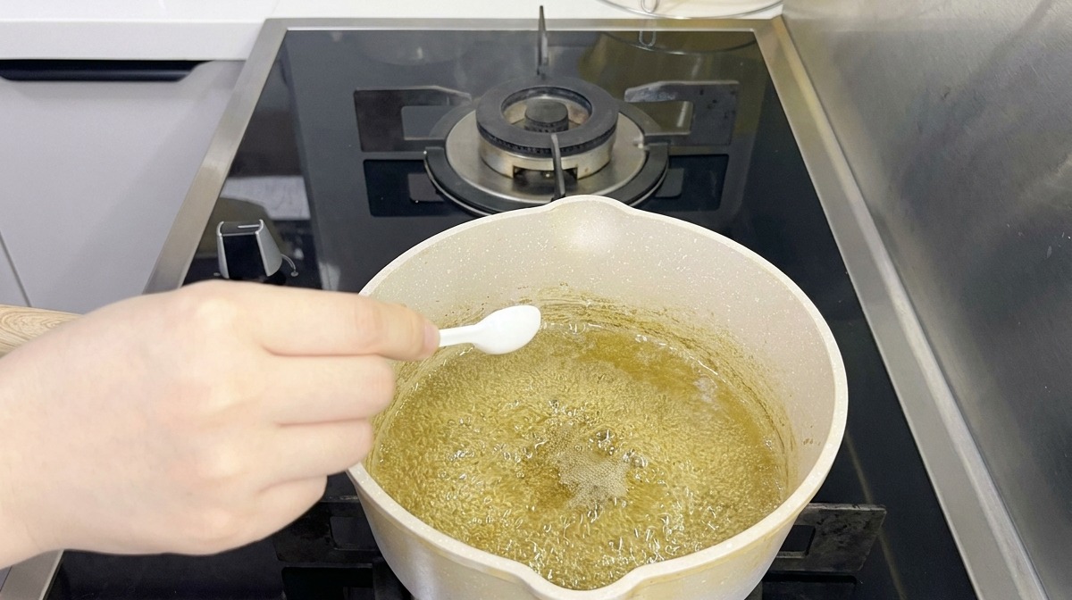 Una persona usa un piccolo cucchiaio bianco per aggiungere condimento a un liquido bollente e verdastro in un pentolino.