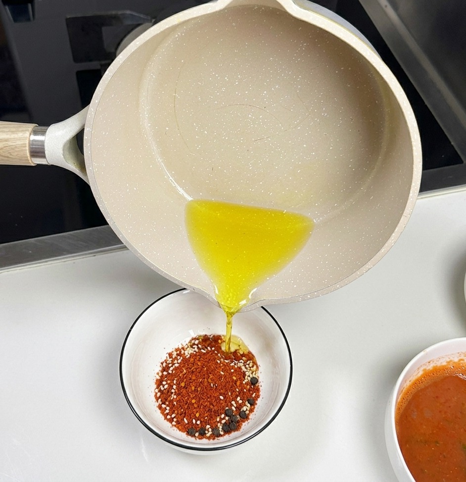 Un pentolino in ceramica chiara versa olio bollente in una ciotolina bianca piena di peperoncino in polvere e semi di sesamo bianchi.