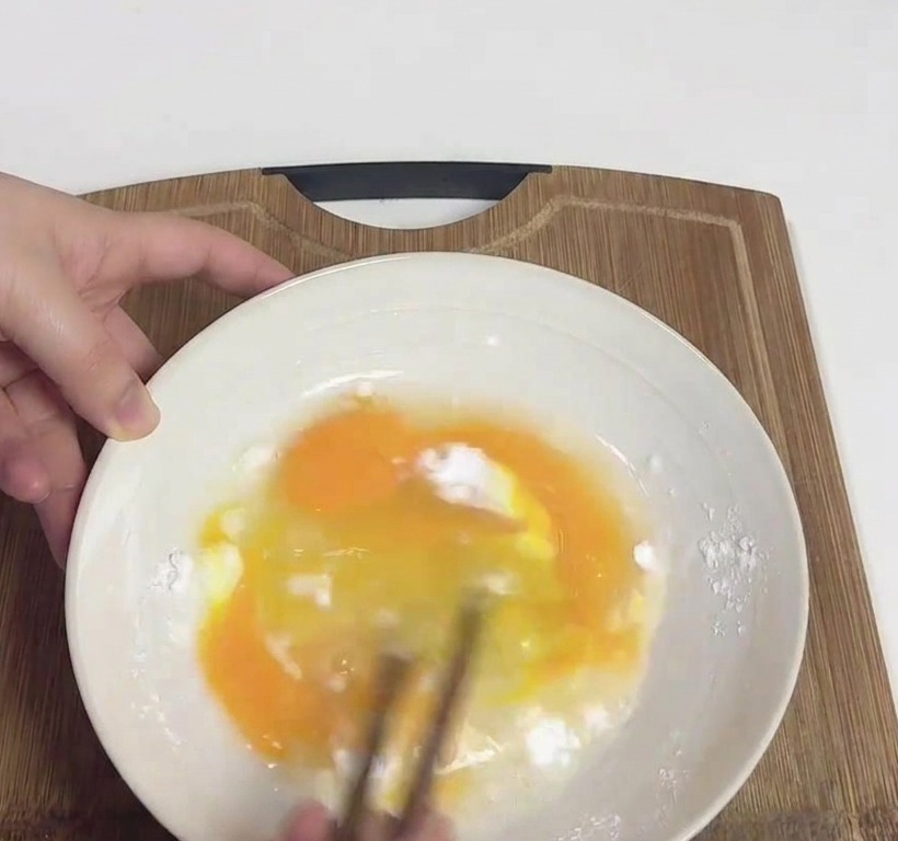 Mani che sbattono uova gialle e fecola di patate bianca in una ciotola usando un paio di bacchette di legno.