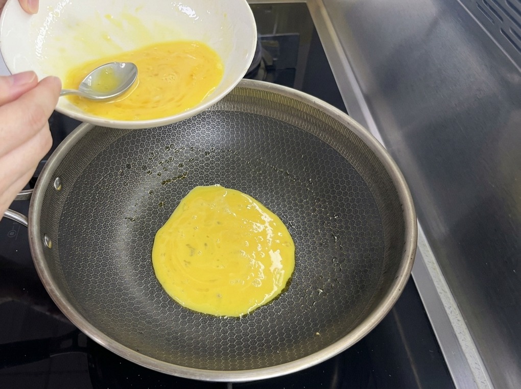 Una sottile pastella di uova viene versata da una ciotola in una grande padella con trama esagonale.