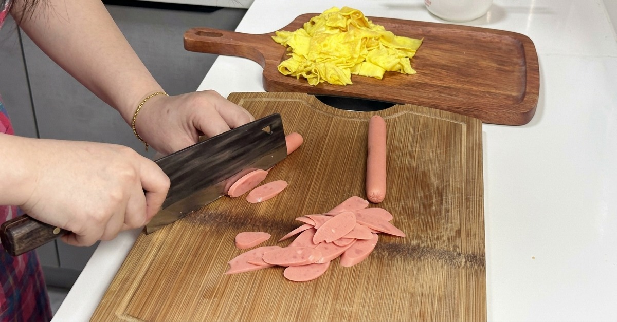 Una persona taglia a fette diagonali dei würstel rosa su un tagliere di legno con una mannaia.