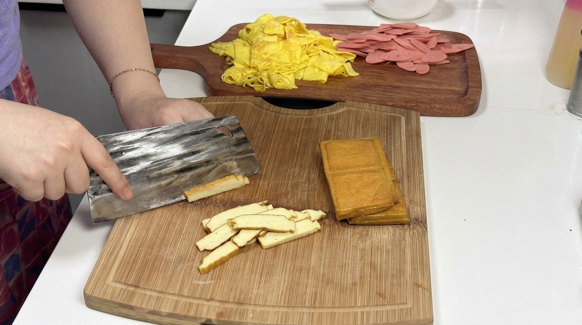 Una mannaia di metallo taglia una pila di panetti di tofu affumicato dorato su un tagliere di legno, con strisce di frittata e prosciutto sullo sfondo.