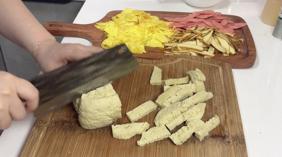 Un grande blocco di glutine al vapore, chiaro e spugnoso, viene tagliato in porzioni rettangolari su un tagliere di legno usando una mannaia.