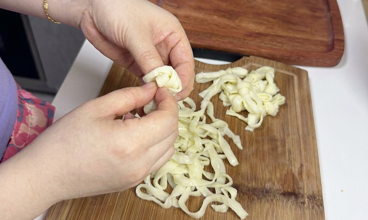 Mani che separano e allentano con cura un mucchio di noodles freddi, larghi e bianchi, su un tagliere di legno.