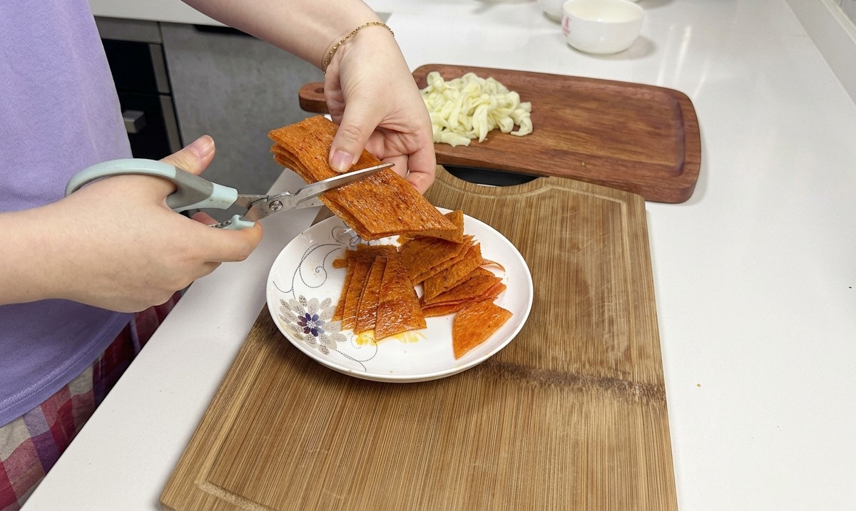 Forbici da cucina tagliano una pila di strisce arancioni di latiao piccante sopra un piatto di ceramica bianca.