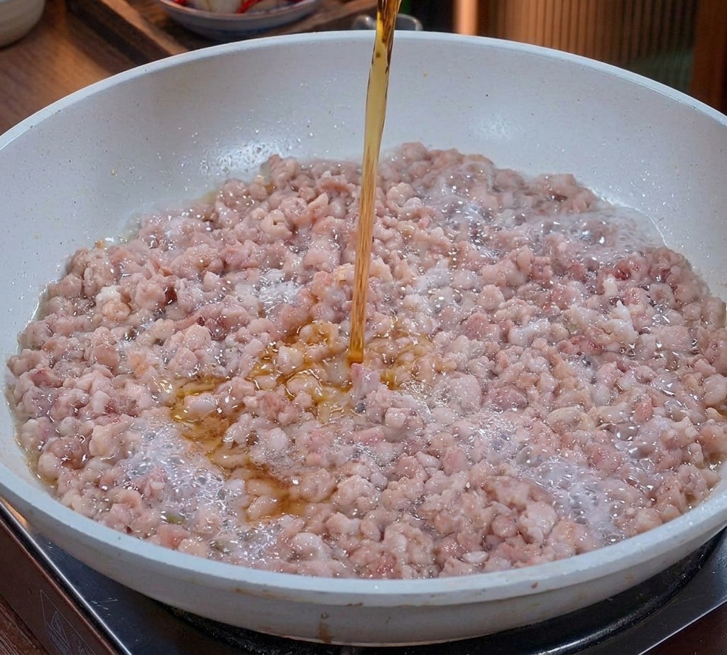 Un liquido ambrato limpido versato da un bicchiere sul macinato di maiale che cuoce in padella.