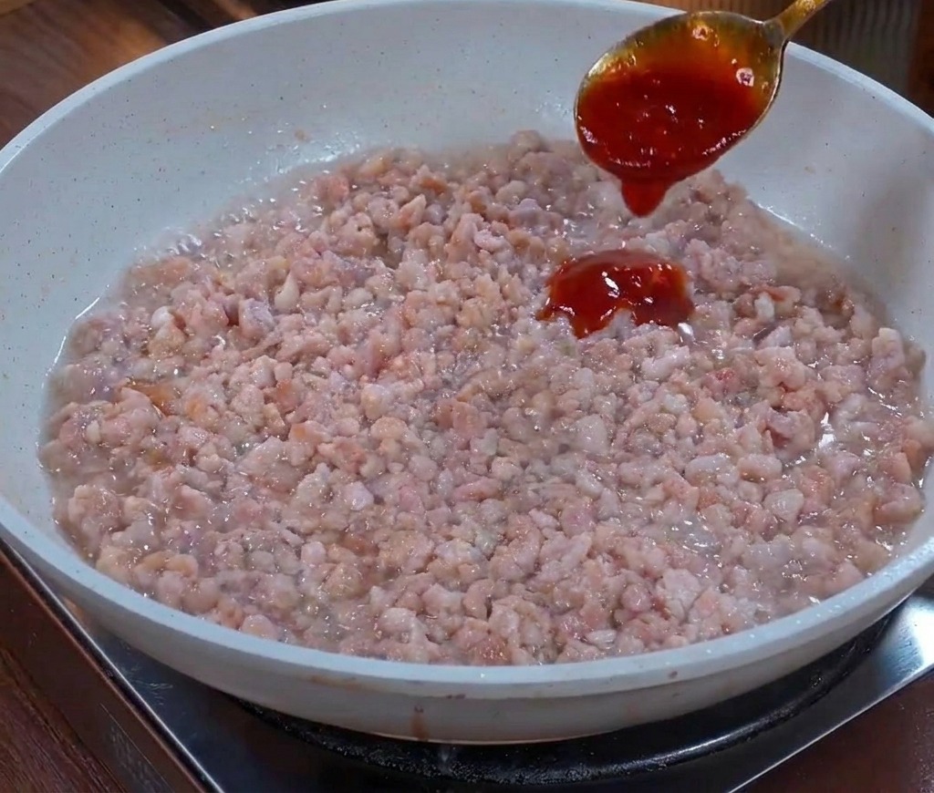 Un cucchiaio di metallo versa una salsa scura e densa su un letto di carne macinata saltata in padella.