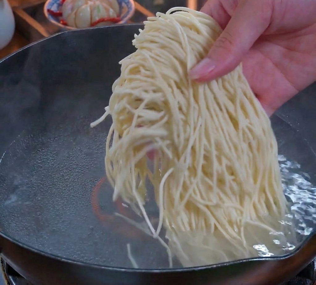 Una mano che cala un nido abbondante di noodles freschi e sottili in una pentola di acqua bollente.