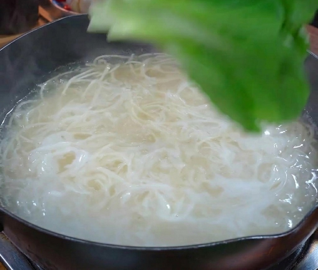 Verdure fresche a foglia verde aggiunte a una pentola dacqua bollente insieme ai noodles bianchi.