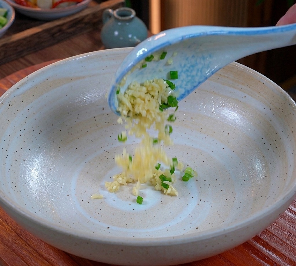 Aglio tritato e cipollotto sminuzzato che cadono da un cucchiaio bianco e blu sul fondo di unampia ciotola di ceramica.