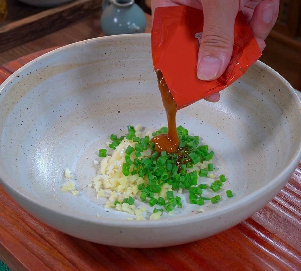Una mano che versa la densa salsa Dandan marrone da una bustina rossa in una ciotola di ceramica sopra laglio e i cipollotti tritatati.