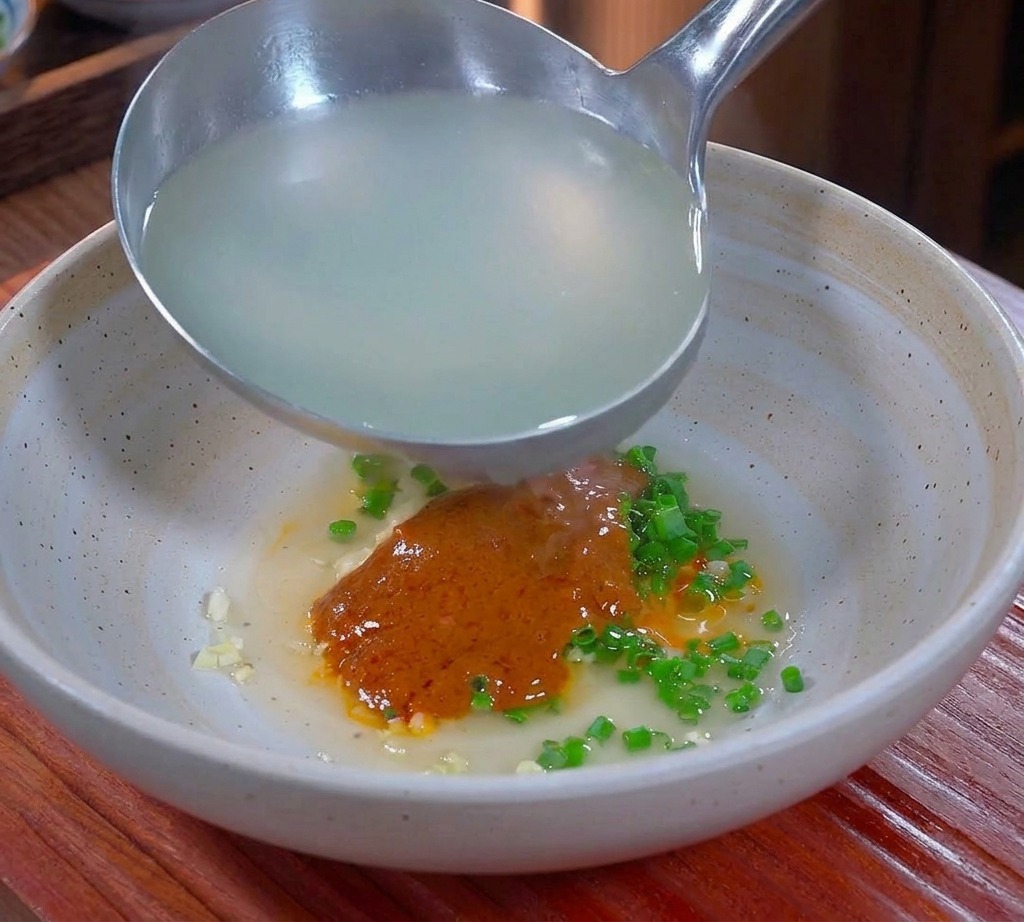 Acqua di cottura dei noodles calda e opaca versata con un mestolo in una ciotola contenente i condimenti piccanti e il grasso di maiale.