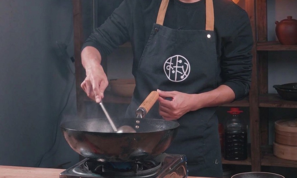 Uno chef salta il maiale macinato in un tradizionale wok nero aggiungendo condimenti liquidi durante la cottura.
