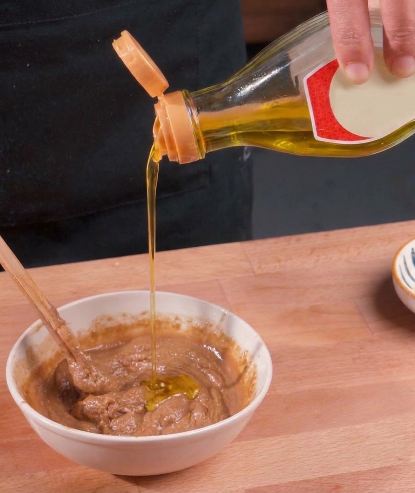 Olio dorato e limpido viene versato da una bottiglia in una ciotola bianca contenente una densa salsa di sesamo scura, mentre viene mescolata con una bacchetta.