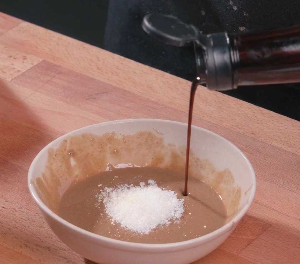 Un filo di salsa di soia scura viene versato in una ciotola di ceramica bianca contenente pasta di sesamo densa, una montagnetta di zucchero bianco e aceto.