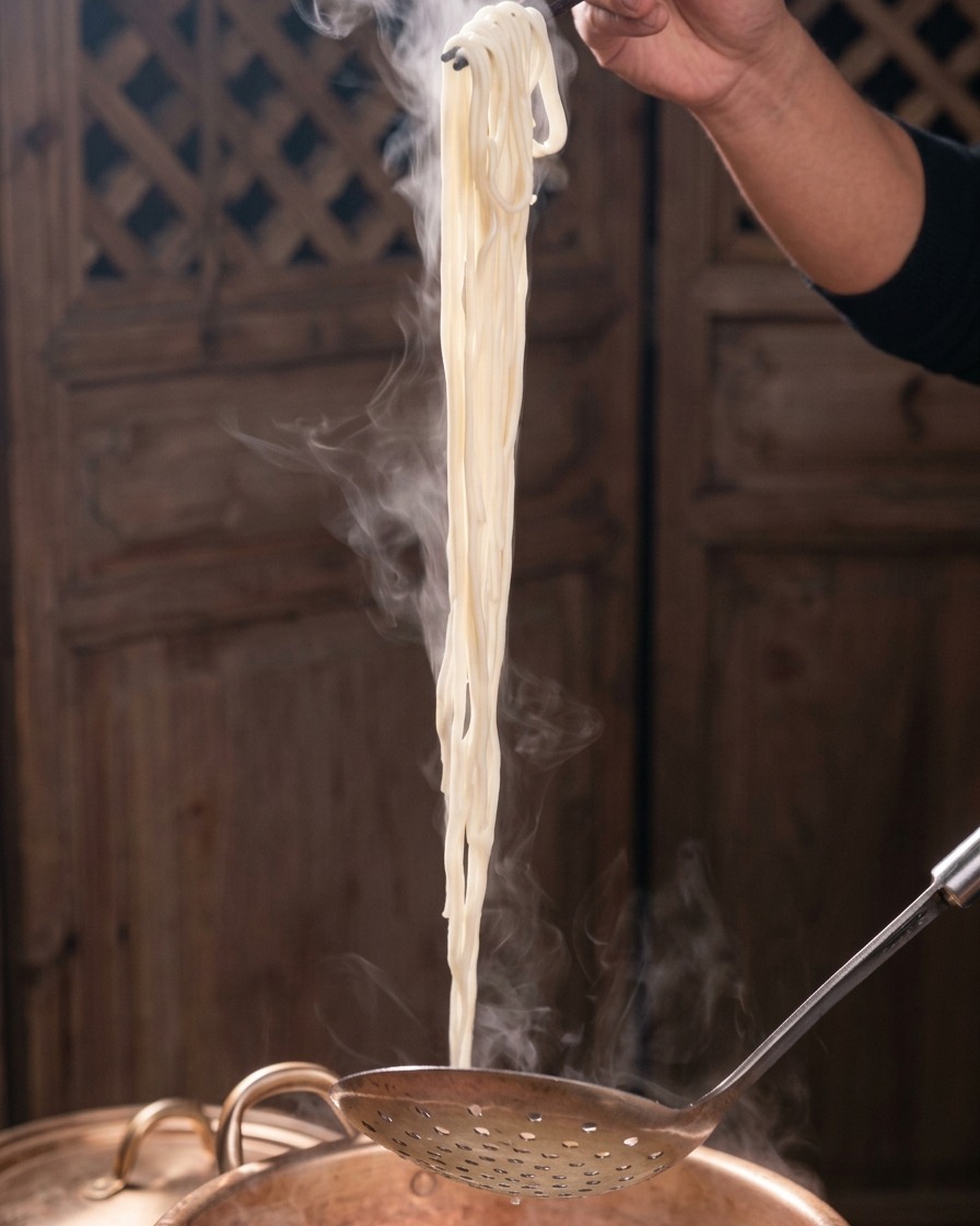 Una matassa di noodles bianchi fumanti viene sollevata verticalmente da una pentola di rame usando bacchette scure e un mestolo forato di metallo.