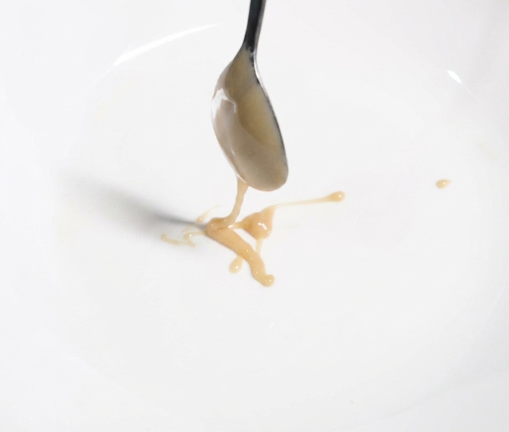 Un cucchiaio di metallo che versa una densa pasta di sesamo marrone chiaro in una ciotola bianca, per creare la base della salsa.