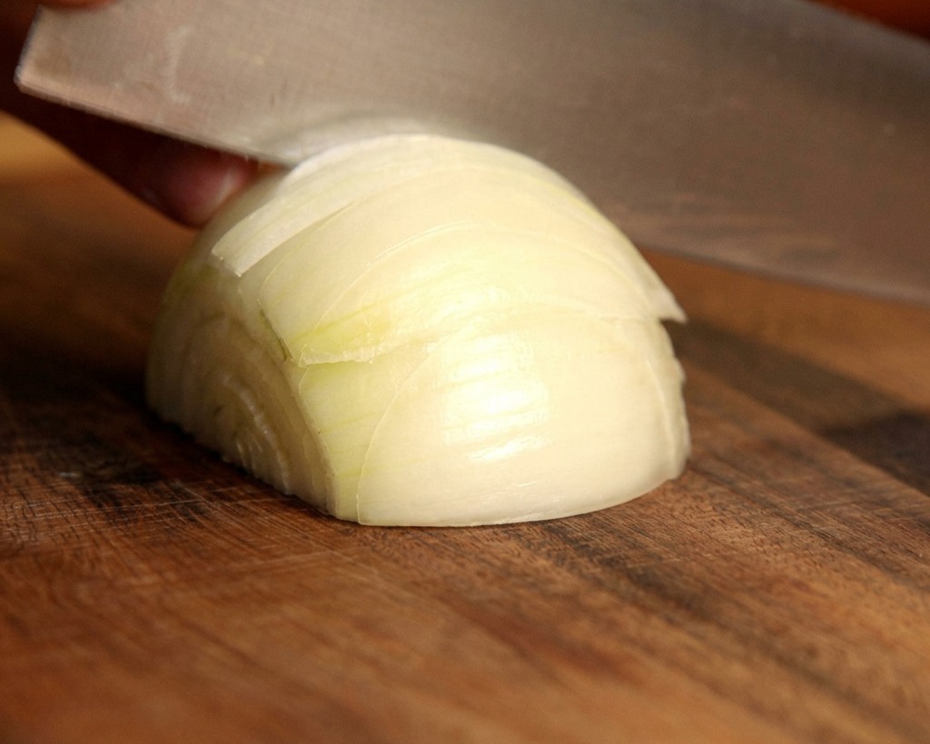 Un coltello affilato taglia una cipolla bianca fresca a spicchi sottili su un tagliere di legno.