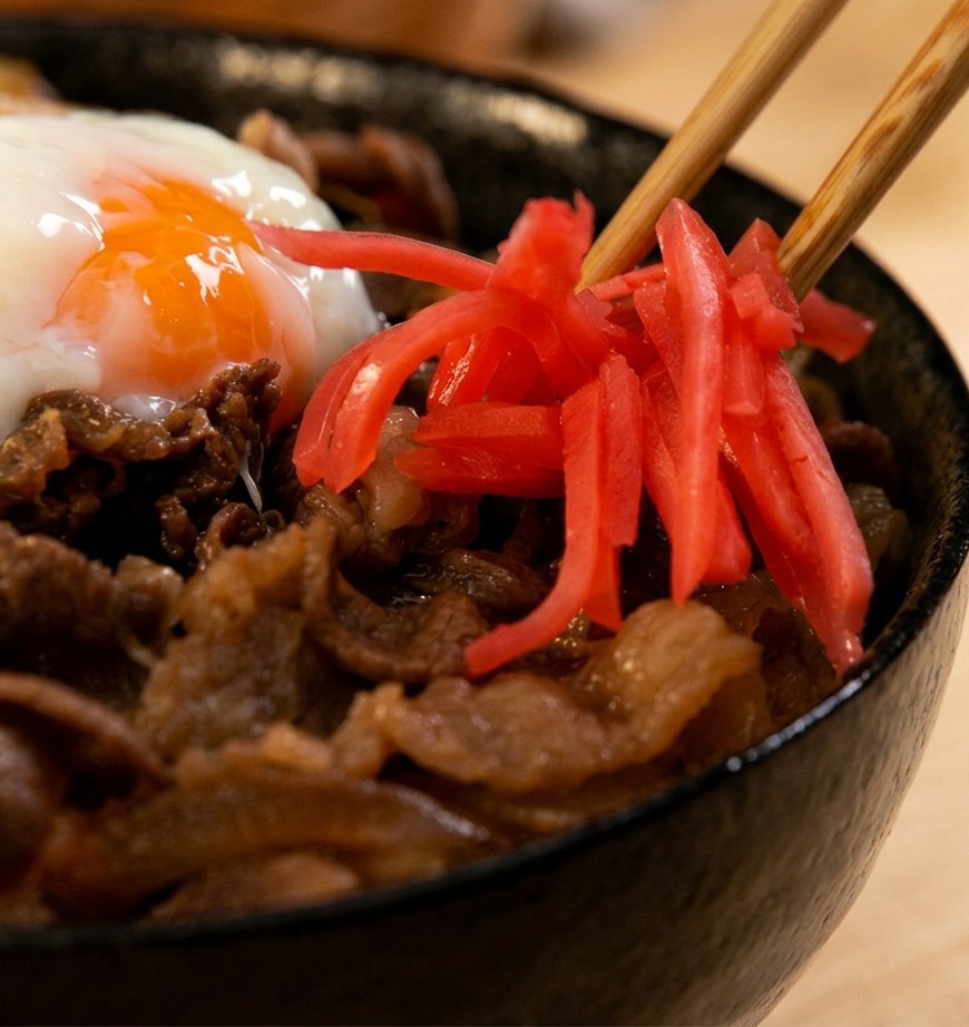 Dettaglio di bacchette di legno che adagiano filetti di zenzero rosso acceso su una ciotola di gyudon con uovo morbido.