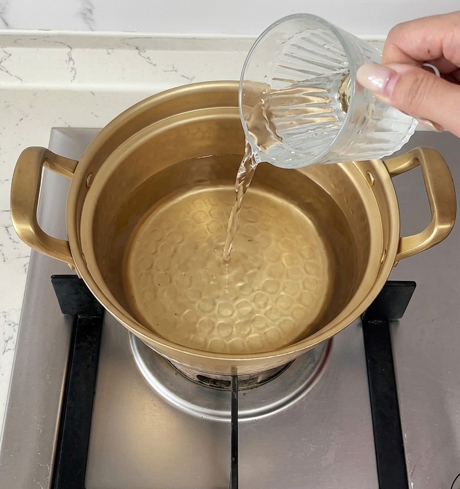 Una mano versa una tazza di acqua fredda in un pentolino dorato sul fuoco contenente acqua già bollente.