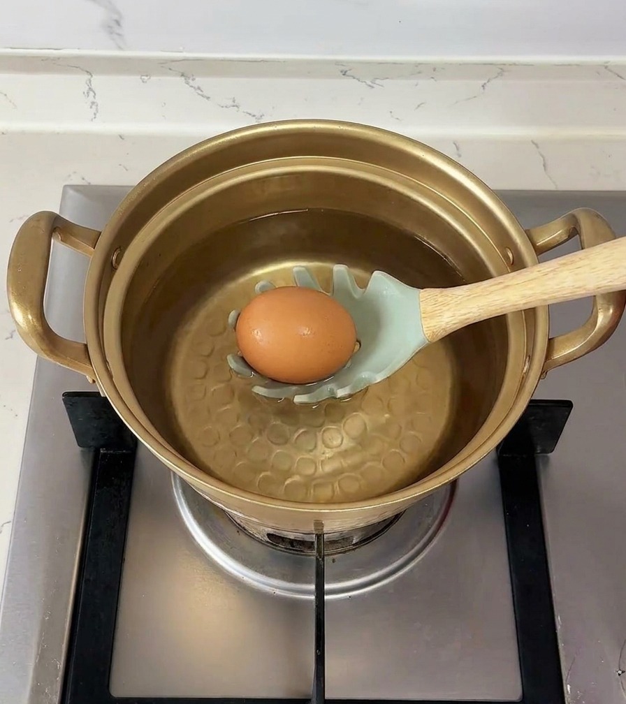 Un uovo marrone adagiato su un cucchiaio forato verde chiaro viene immerso con cura nel pentolino dacqua.
