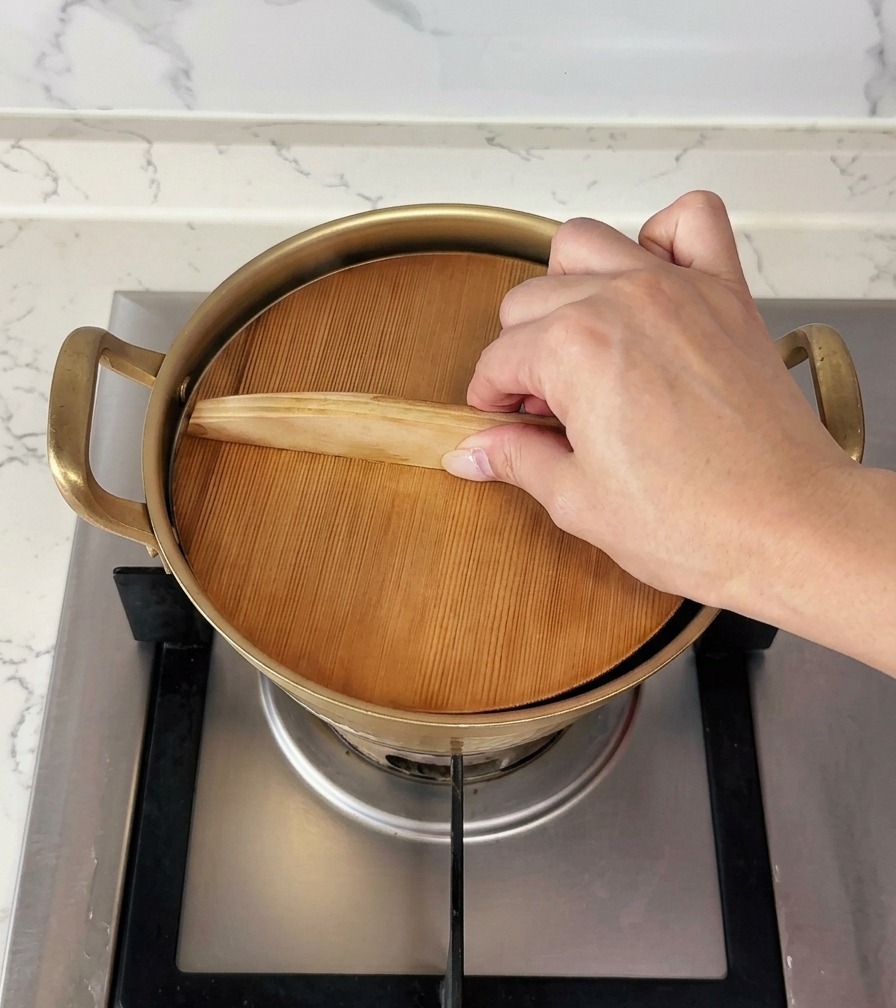 Una mano posiziona un coperchio piatto di legno per coprire un piccolo pentolino dorato.