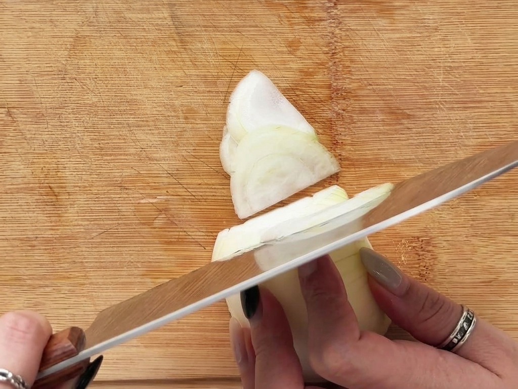 Mani che affettano una cipolla bianca a mezzelune sottili su un tagliere di legno.