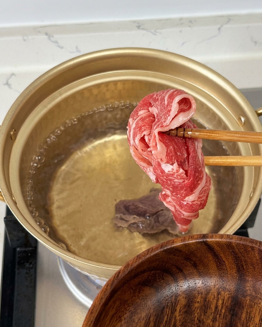 Una sottile fettina di manzo crudo e marmorizzato tenuta con le bacchette sopra un pentolino dorato di acqua bollente.