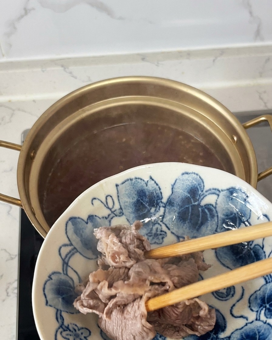 Fettine di manzo parzialmente cotte e opache sollevate dal pentolino con le bacchette e trasferite su un piatto decorato bianco e blu.