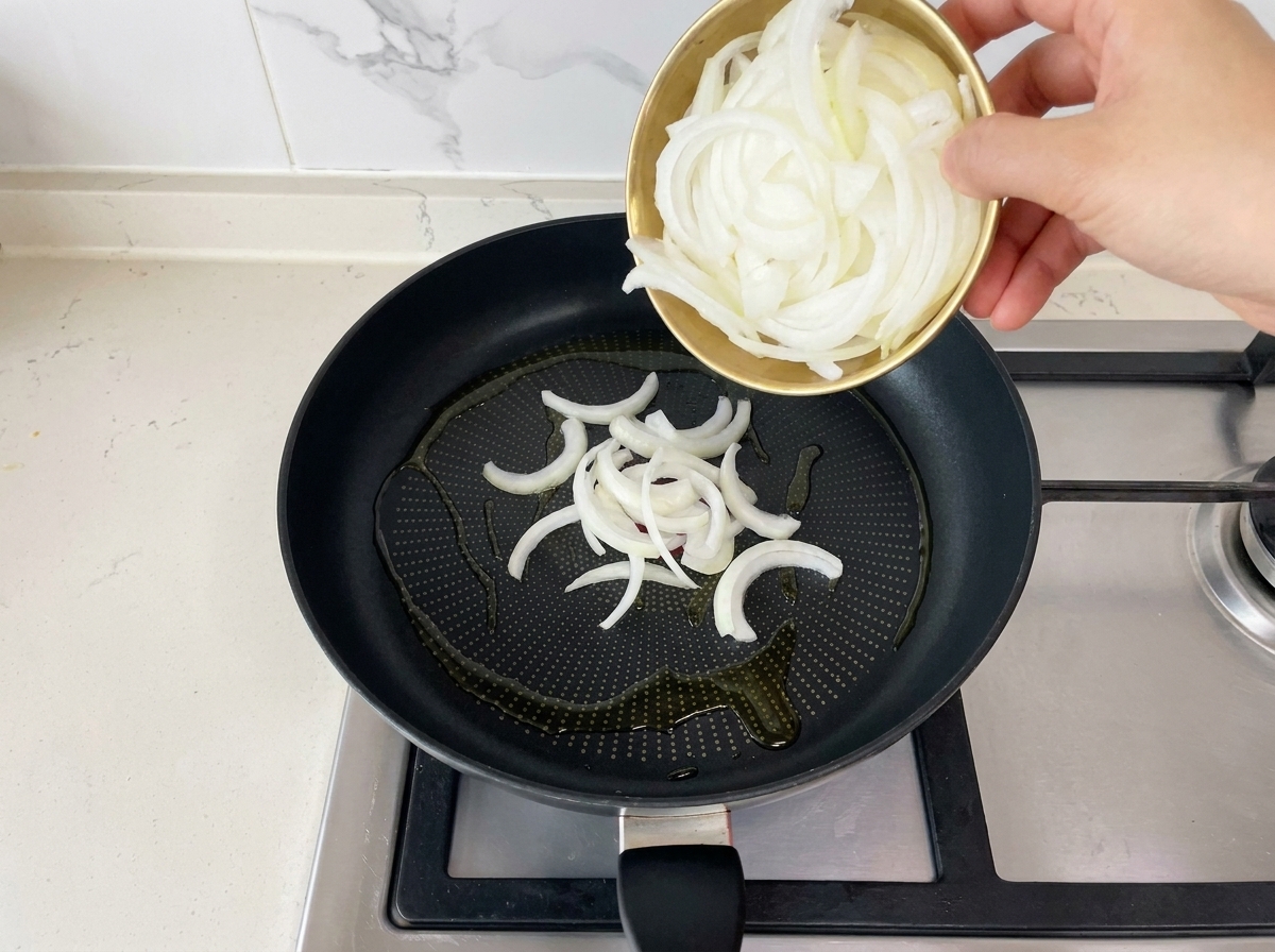 Cipolle bianche appena affettate versate da una ciotolina dorata in una padella nera con olio caldo.