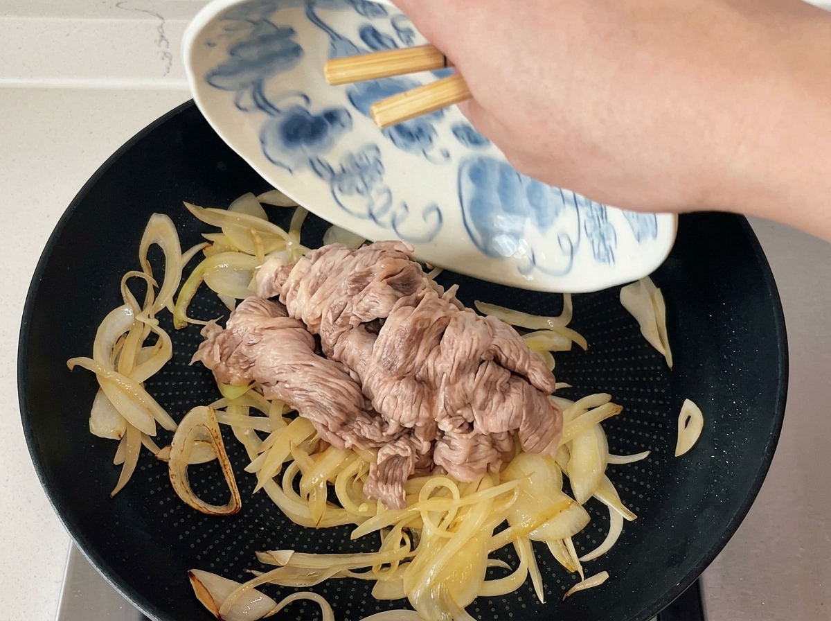 Bacchette che fanno scivolare le fettine di manzo sbollentate da un piatto decorato a una padella con cipolle saltate.