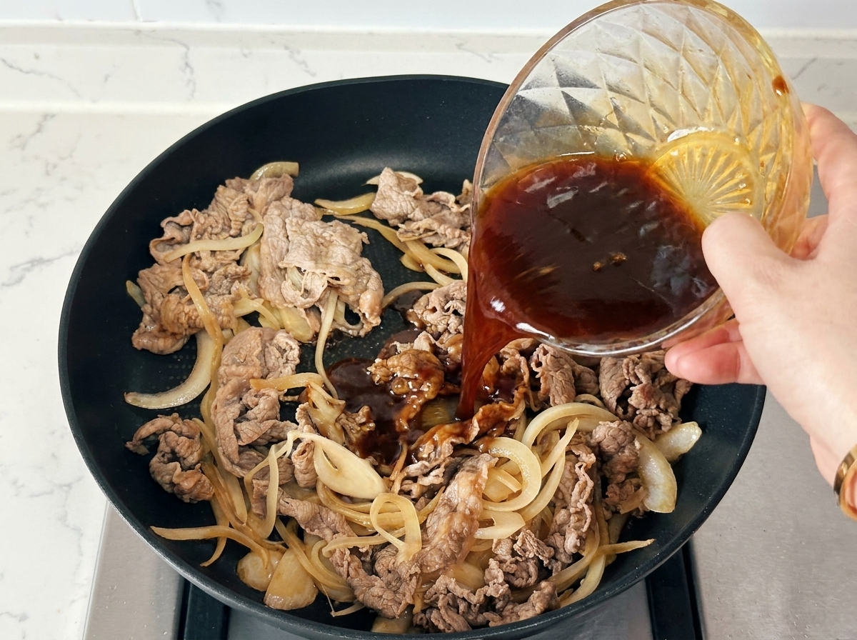 Mano che versa una salsa scura da una ciotola di vetro sopra il manzo e le cipolle in una padella nera.