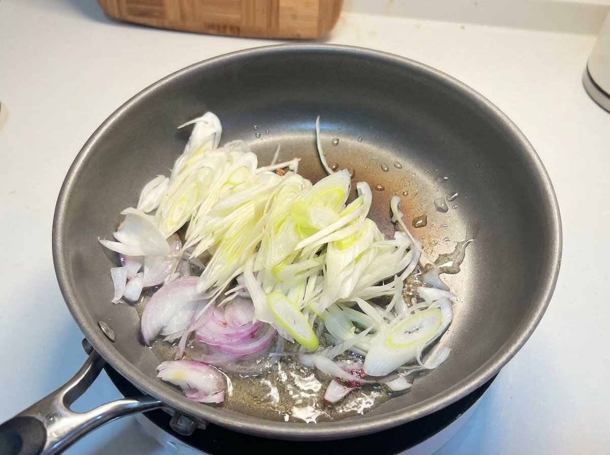 Cipolle bianche affettate e cipollotti che sfrigolano in olio caldo dentro una padella di metallo.