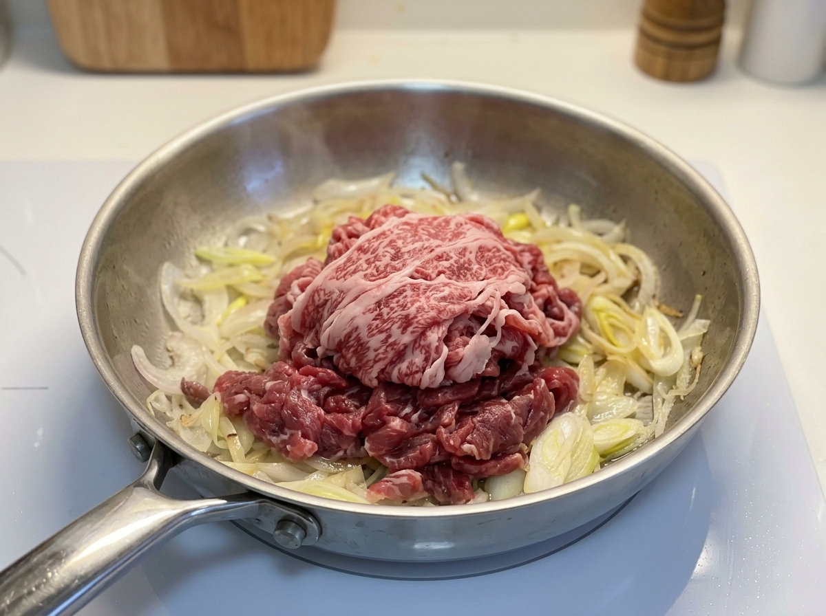 Una porzione di carne di manzo cruda a fettine sottili adagiata sopra le cipolle cotte in una padella di metallo.