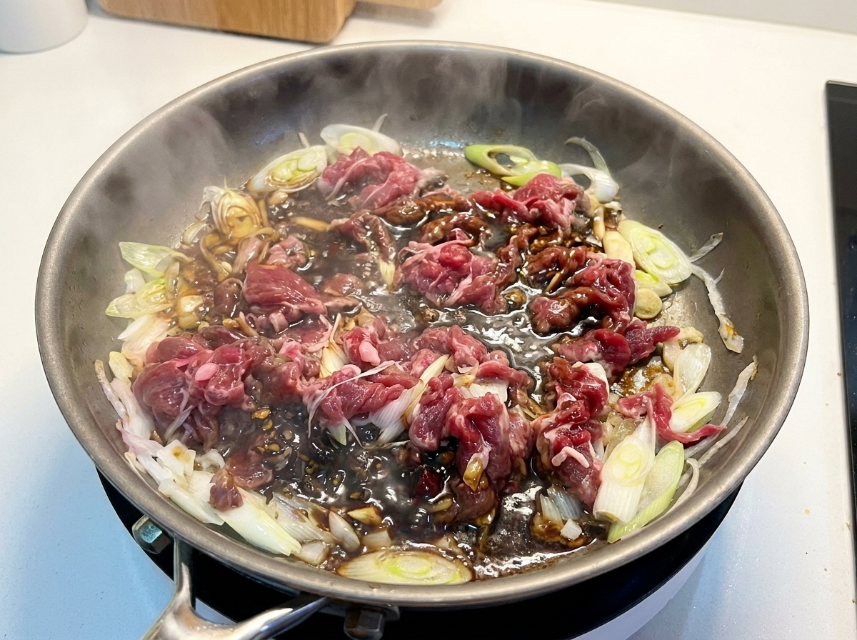 Fettine di manzo parzialmente cotte e cipolle immerse in una salsa scura e ricca che sobbolle in padella.
