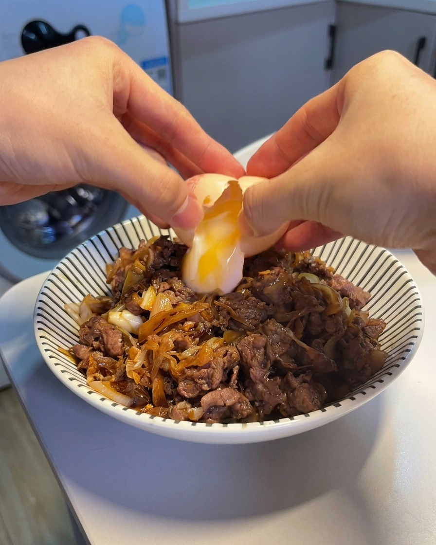 Due mani che rompono un uovo onsen, facendo cadere il tuorlo morbido e lalbume su una ciotola fumante di manzo e riso.