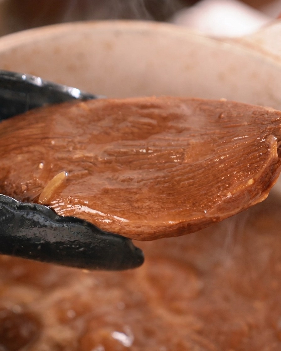 Un pezzo spesso di lingua di manzo brasata viene sollevato da una pentola con salsa scura usando pinze da cucina nere.