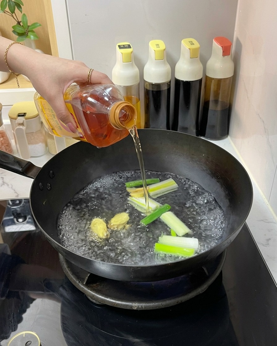 Una persona che versa un liquido ambrato da una bottiglia di plastica in un wok con acqua bollente, zenzero e cipollotti.