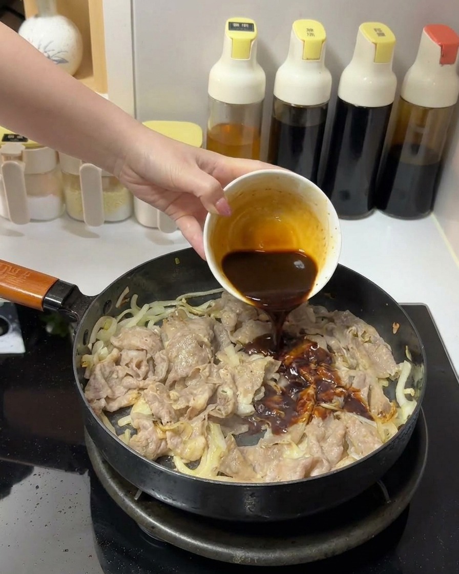 Una salsa densa e marrone scuro versata da una ciotolina bianca su manzo e cipolle in padella.
