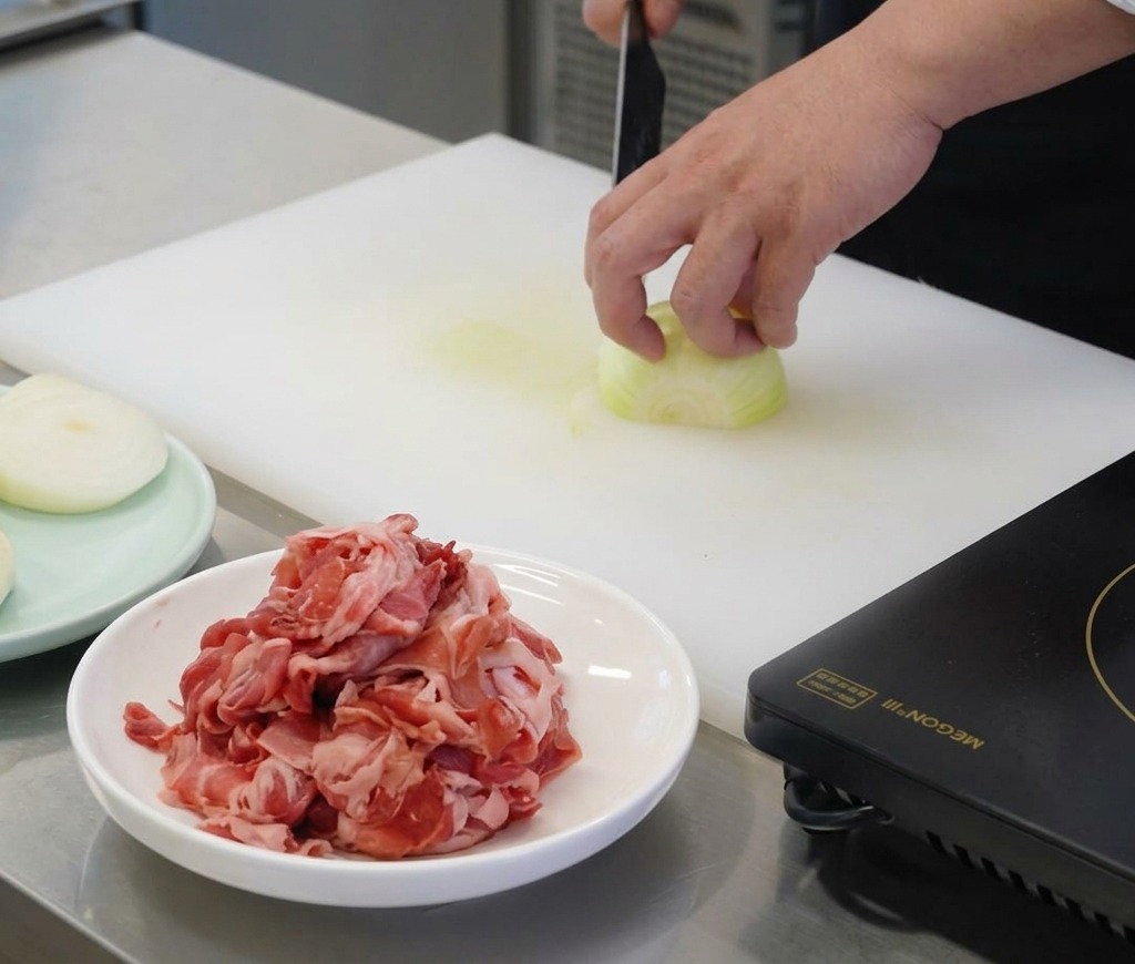 Mano di uno chef che tiene una cipolla bianca su un tagliere bianco mentre la taglia a fette sottili verticali con un coltello affilato.