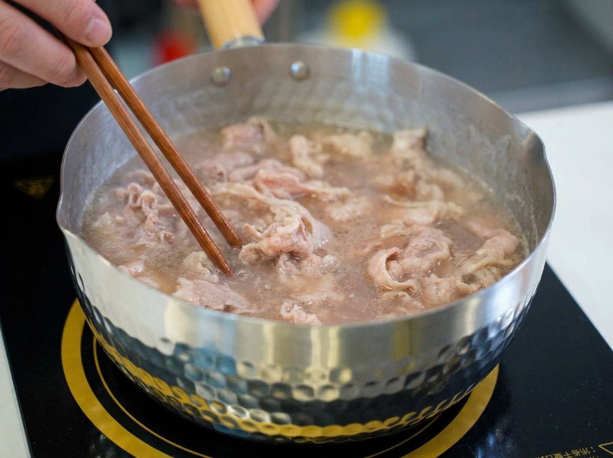 Sottili fettine di manzo mescolate con bacchette di legno in una pentola di acqua che sobbolle mentre virano dal rosa crudo al marrone chiaro.