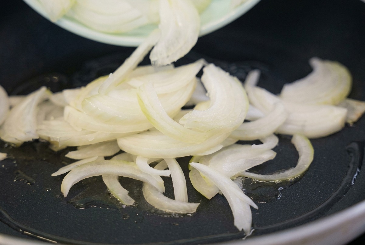 Fettine di cipolla bianca cruda versate da un piatto verde chiaro in una padella nera con olio di cottura caldo.