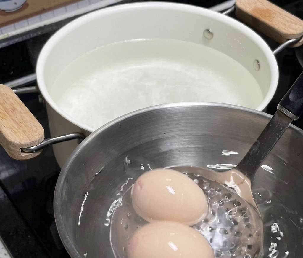 Due uova in una ciotola di acciaio inossidabile piena di acqua e ghiaccio, con la pentola bianca usata per la cottura visibile sullo sfondo.